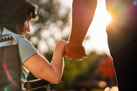 Close-up of a caregiver holding hands with a woman sitting in a wheelchair outdoors during a warm golden hour sunset.