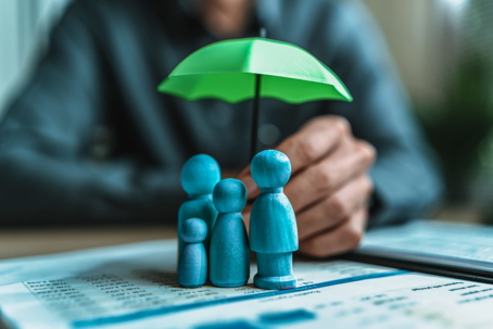 A person holding a small green umbrella over a family of wooden figures resting on financial documents, symbolizing family protection through a life insurance trust.
