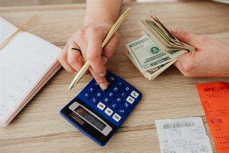 Hands using a calculator while holding cash and a pen, with receipts and a planner on a desk, suggesting budgeting or expense tracking.