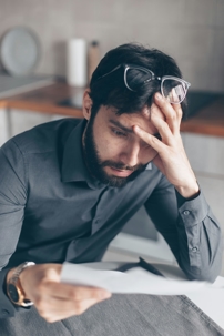 A man sits at a table holding a piece of paper while resting his head on his hand, appearing stressed or concerned as he reads the document.