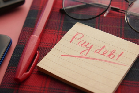 A sticky note with the handwritten words “Pay debt” underlined in red sits on a desk next to a red pen, glasses, and a phone.