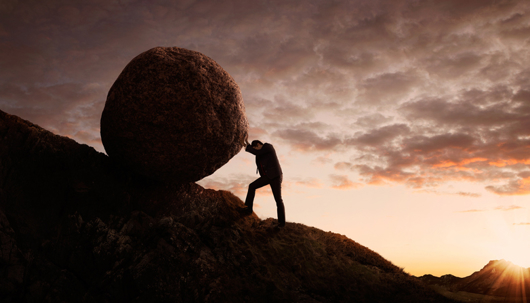 Man pushing a large boulder uphill at sunset symbolizing financial struggle and bankruptcy challenges.
